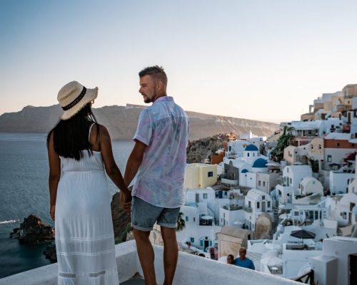 Santorini Greece, young couple on luxury vacation at the Island of Santorini watching sunrise by the blue dome church and whitewashed village of Oia Santorini Greece during sunrise during summer vacation, men and woman on holiday in Greece